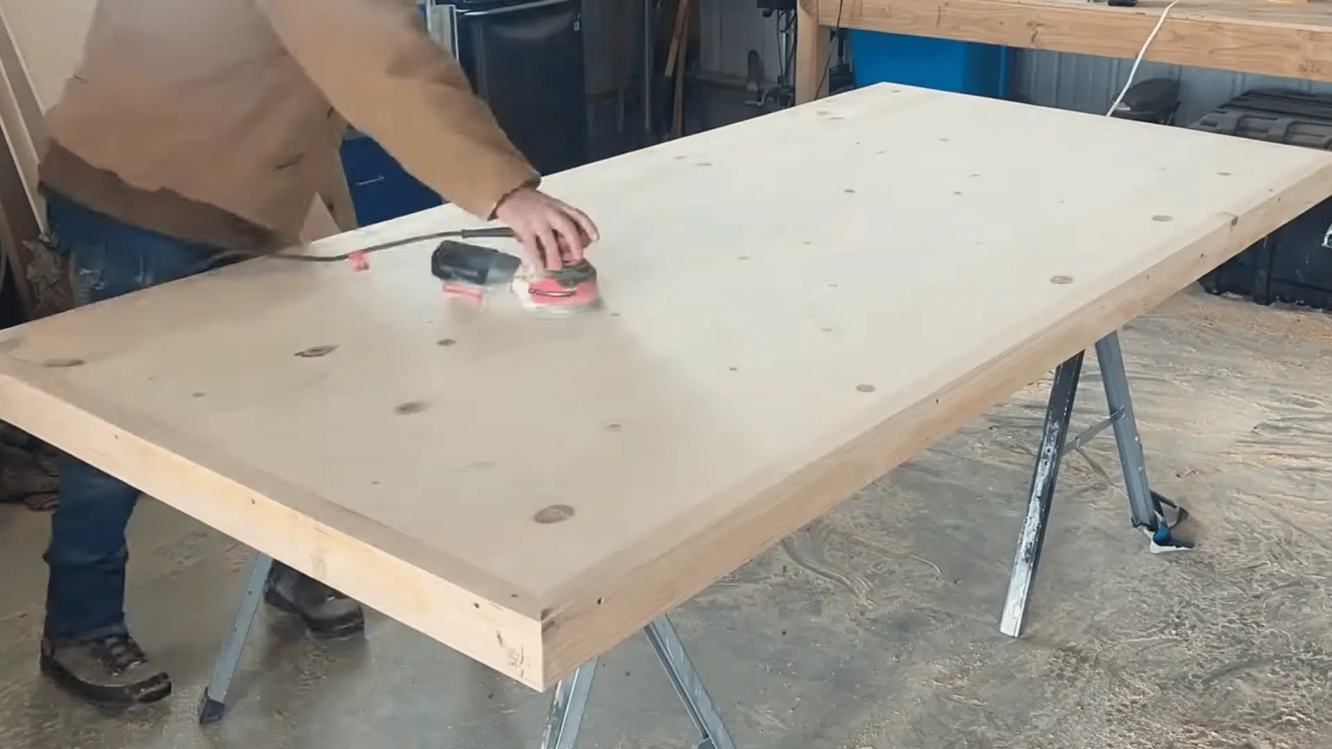 a woodworker sanding a large wooden tabletop with a power sander to smooth the surface before finishing