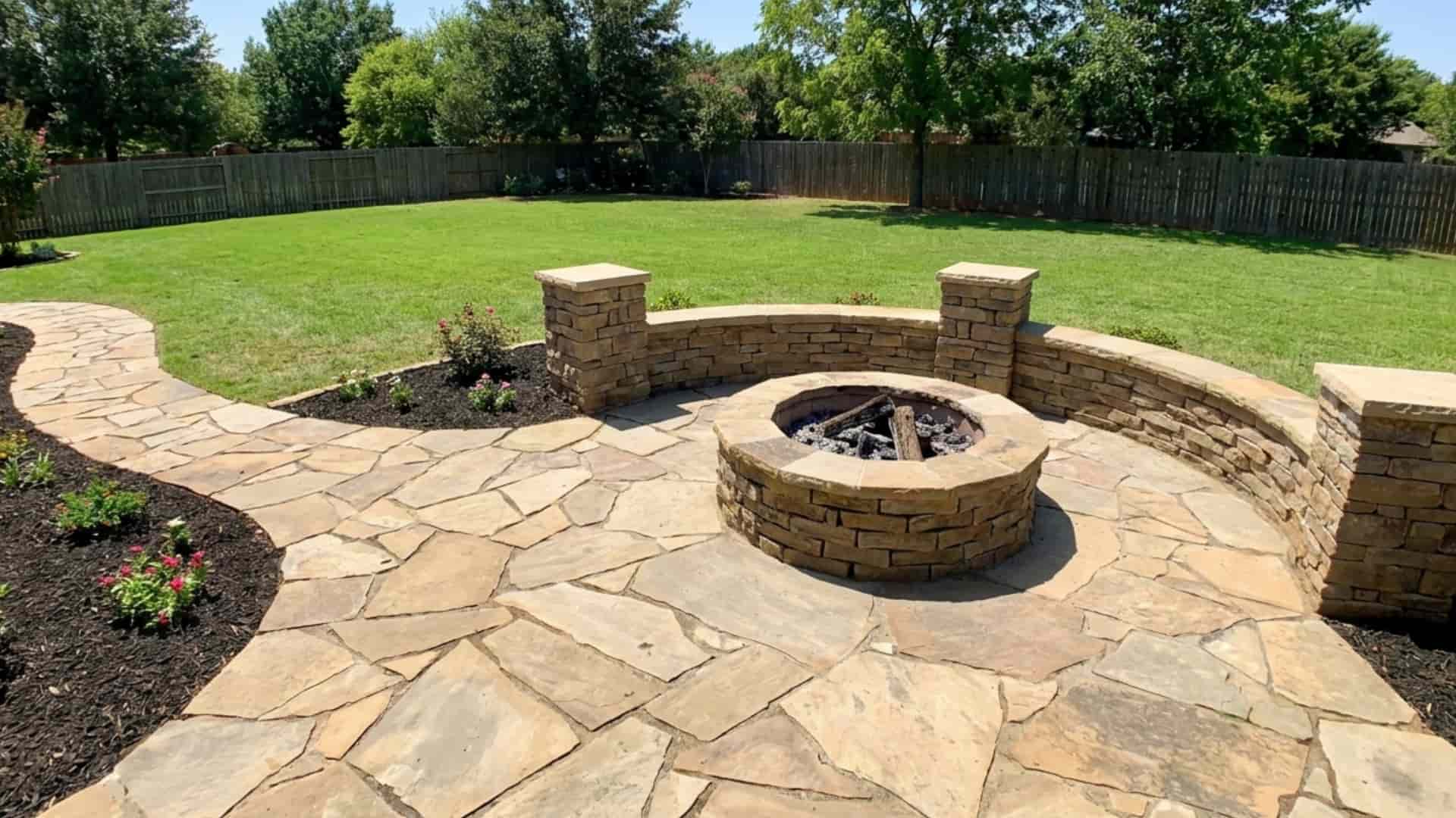 Backyard flagstone patio with curved stone seating wall, circular DIY fire pit, landscaped garden beds, and large green lawn in daylight.
