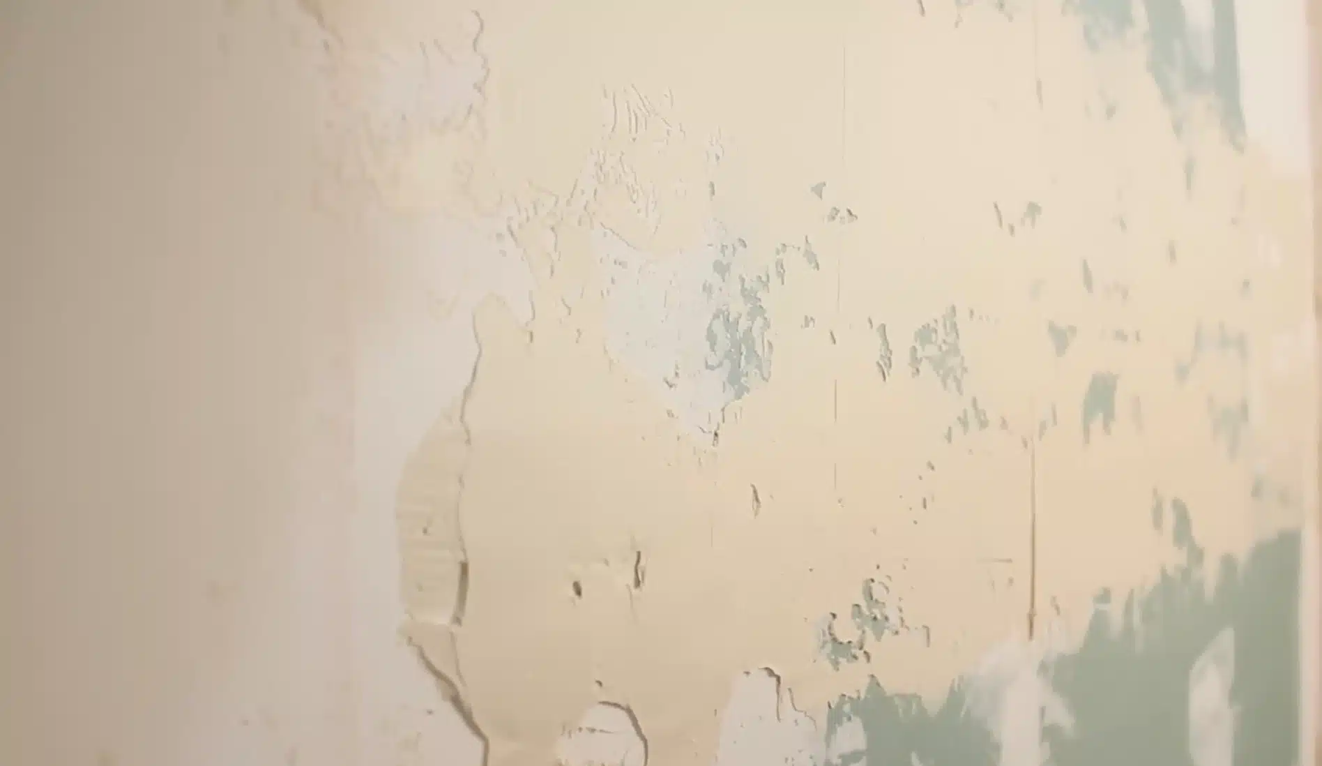 close up of a wall surface where pale yellow paint or drywall compound is peeling away, revealing patches of light blue or green underlayer