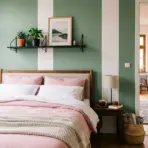 Cozy bedroom featuring a wooden bed with pink and white linens against a sage green and white vertically striped wall, opening into a hallway with light pink walls