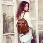 Woman with brown leather backpack standing in front of large windows indoors