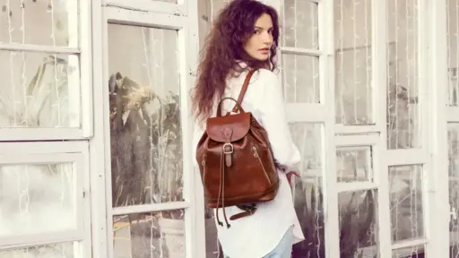 Woman with brown leather backpack standing in front of large windows indoors