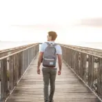 Man with backpack walking on wooden bridge towards sunset over the ocean