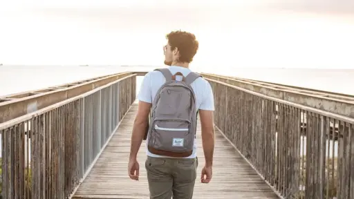 Man with backpack walking on wooden bridge towards sunset over the ocean