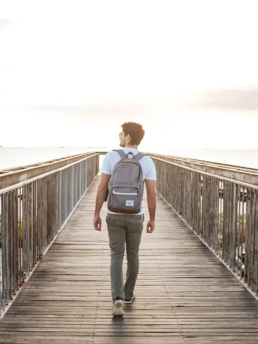 Man with backpack walking on wooden bridge towards sunset over the ocean