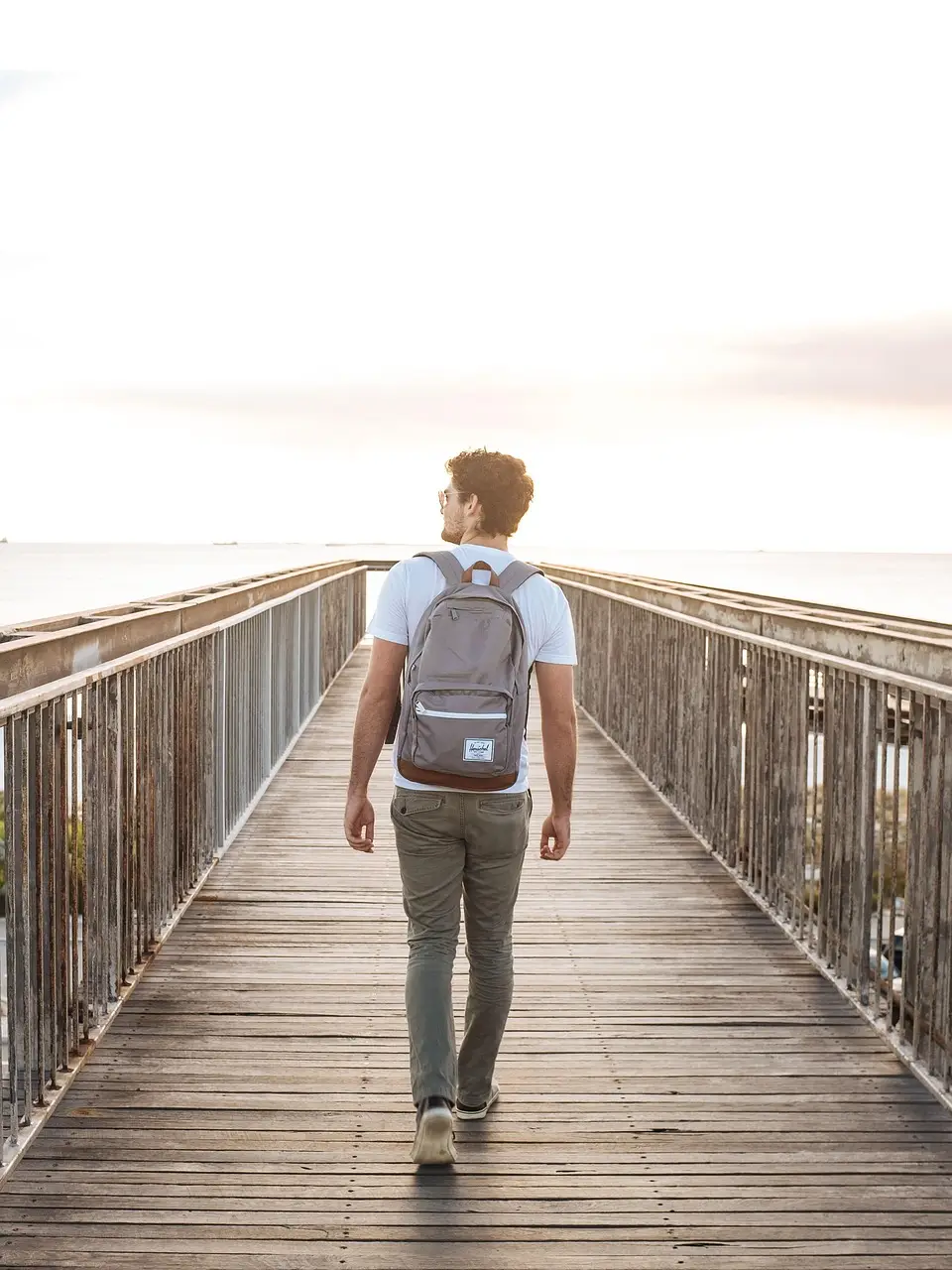 Man with backpack walking on wooden bridge towards sunset over the ocean