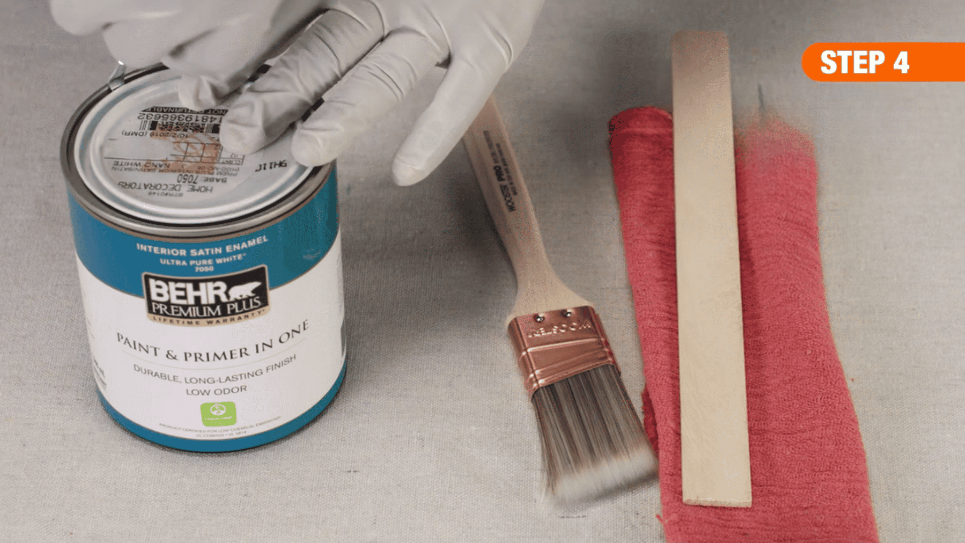 gloved hands opening a can of paint and primer beside a brush sanding stick and cloth for wood refinishing preparation