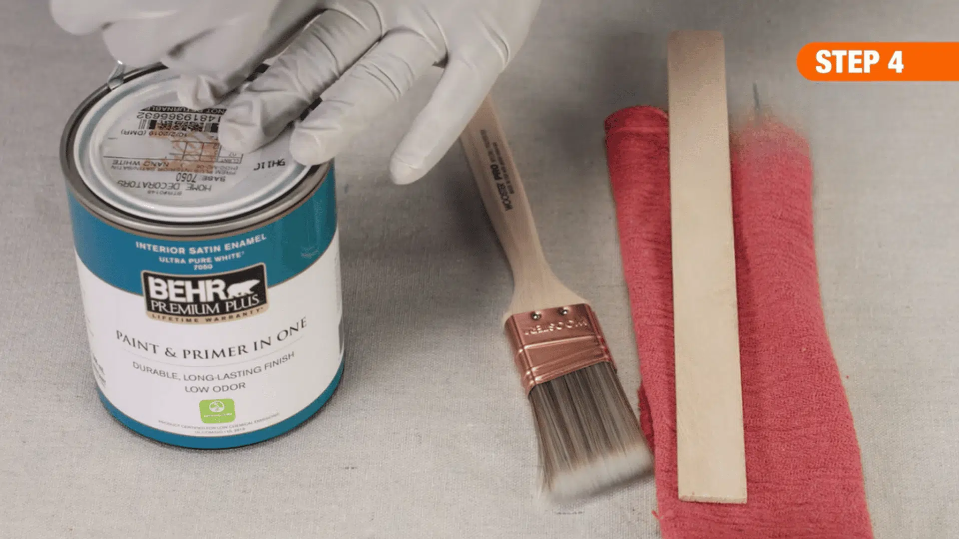 gloved hands opening a can of paint and primer beside a brush sanding stick and cloth for wood refinishing preparation