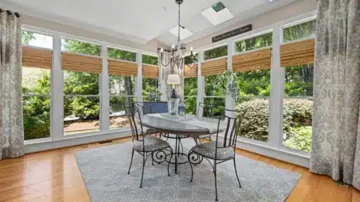 Elegant dining room with glass table, wrought iron chairs, surrounded by large windows and greenery