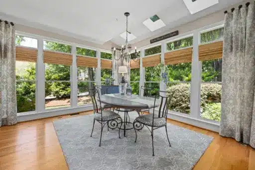 Elegant dining room with glass table, wrought iron chairs, surrounded by large windows and greenery