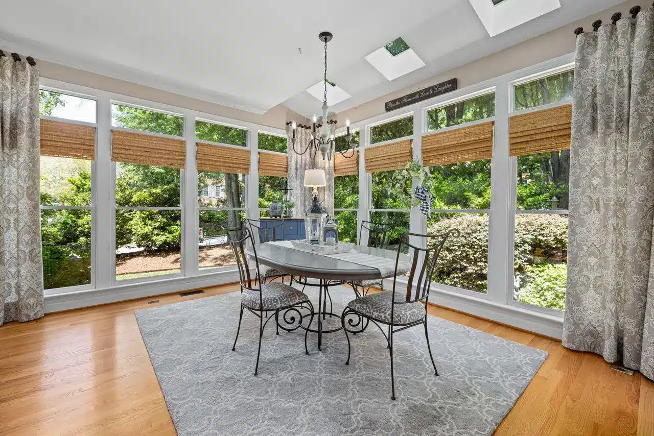 Elegant dining room with glass table, wrought iron chairs, surrounded by large windows and greenery