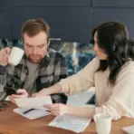 Man holding phone looks shocked as woman points at paperwork on kitchen table