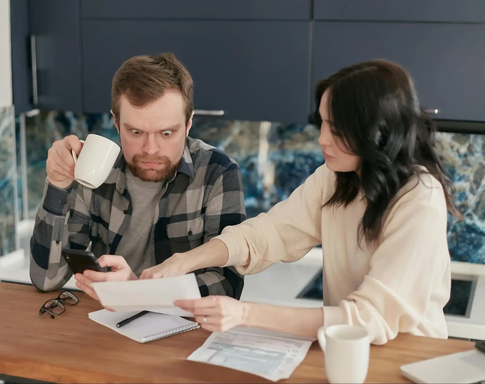 Man holding phone looks shocked as woman points at paperwork on kitchen table
