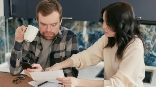 Man holding phone looks shocked as woman points at paperwork on kitchen table