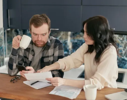 Man holding phone looks shocked as woman points at paperwork on kitchen table