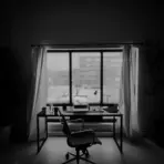 Office desk with laptop and chair next to large window with closed curtains in dim lighting