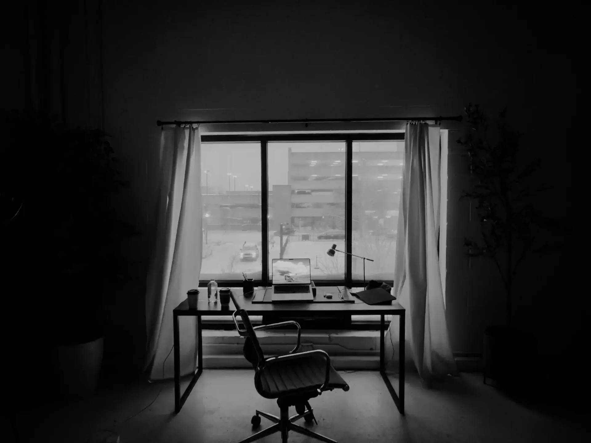 Office desk with laptop and chair next to large window with closed curtains in dim lighting