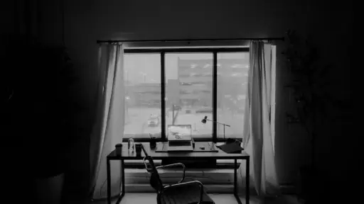 Office desk with laptop and chair next to large window with closed curtains in dim lighting