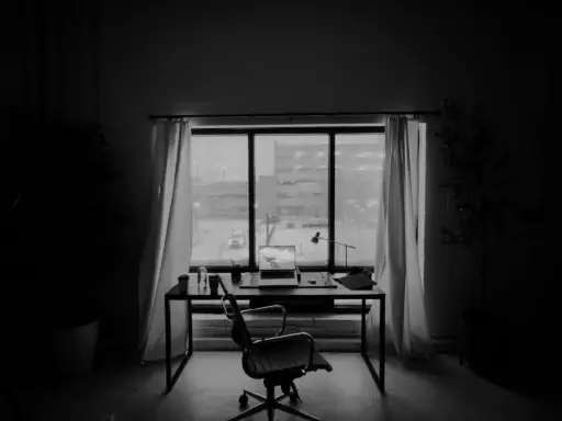 Office desk with laptop and chair next to large window with closed curtains in dim lighting