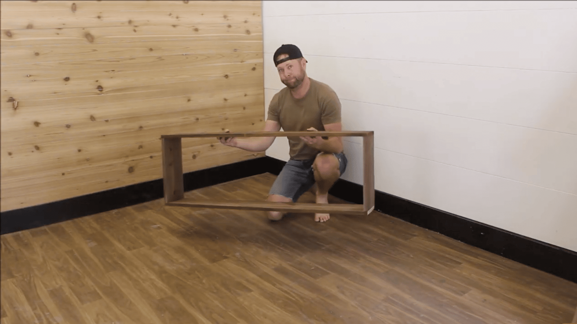 man in cap holding a rectangular wooden frame by his fingers while crouched down on the wooden furnished floor