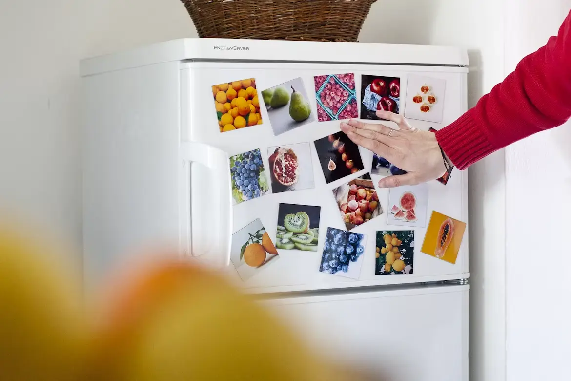 Refrigerator door covered with fruit-themed photo magnets, person placing a hand on the fridge
