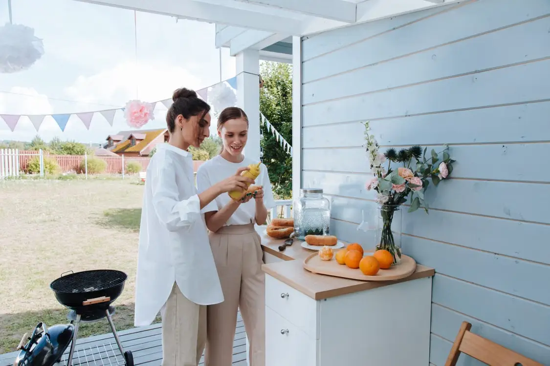 Two people outdoors applying mustard to hot dogs near oranges on a patio table