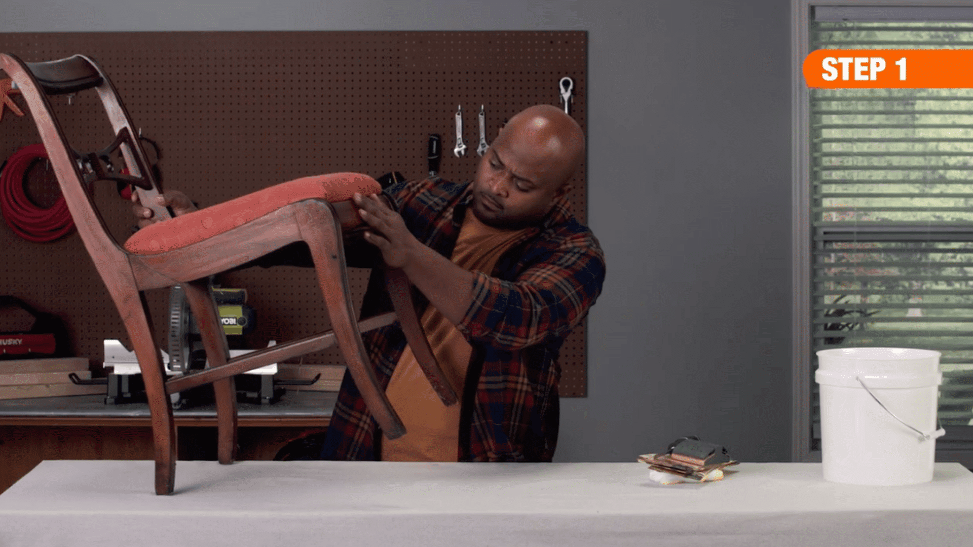 person inspecting and cleaning an upholstered wooden chair on a workbench in a workshop beside sanding tools and bucket
