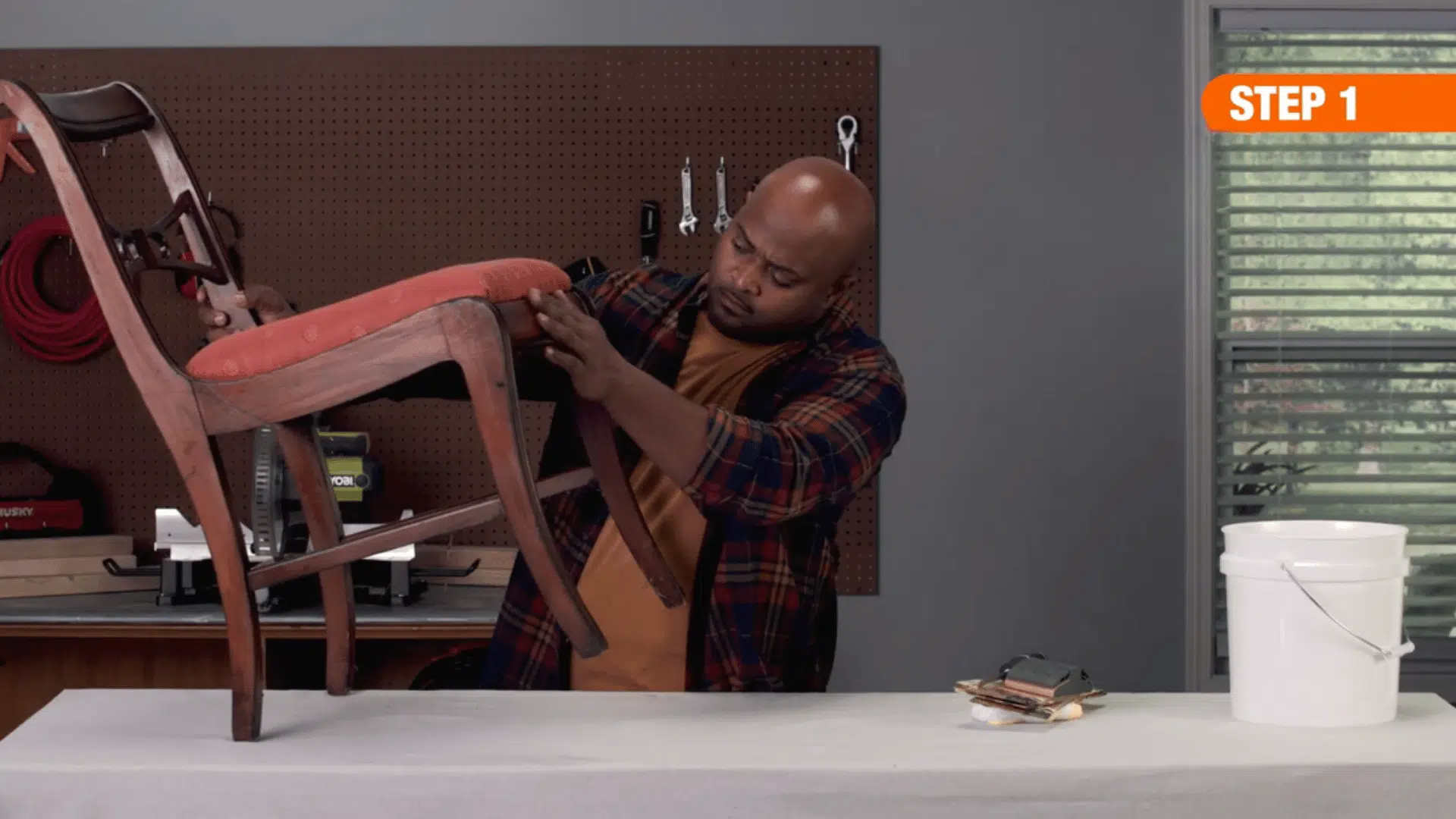 person inspecting and cleaning an upholstered wooden chair on a workbench in a workshop beside sanding tools and bucket