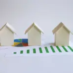 Three miniature wooden houses on financial charts with pie and bar graphs in a bright setting