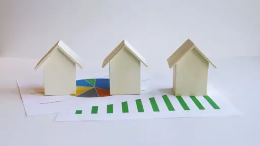 Three miniature wooden houses on financial charts with pie and bar graphs in a bright setting