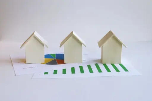 Three miniature wooden houses on financial charts with pie and bar graphs in a bright setting