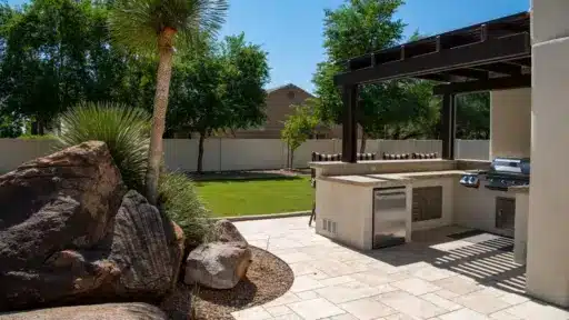 Outdoor kitchen with modern grill and appliances on stone patio under pergola in sunny backyard