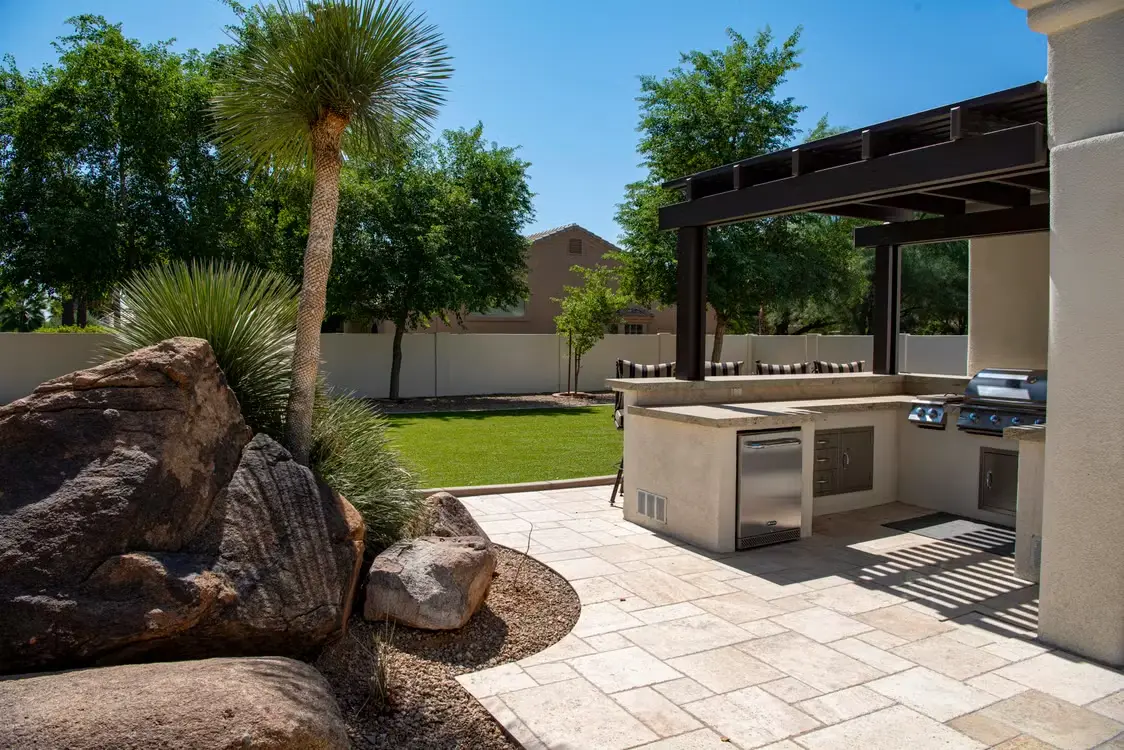Outdoor kitchen with modern grill and appliances on stone patio under pergola in sunny backyard