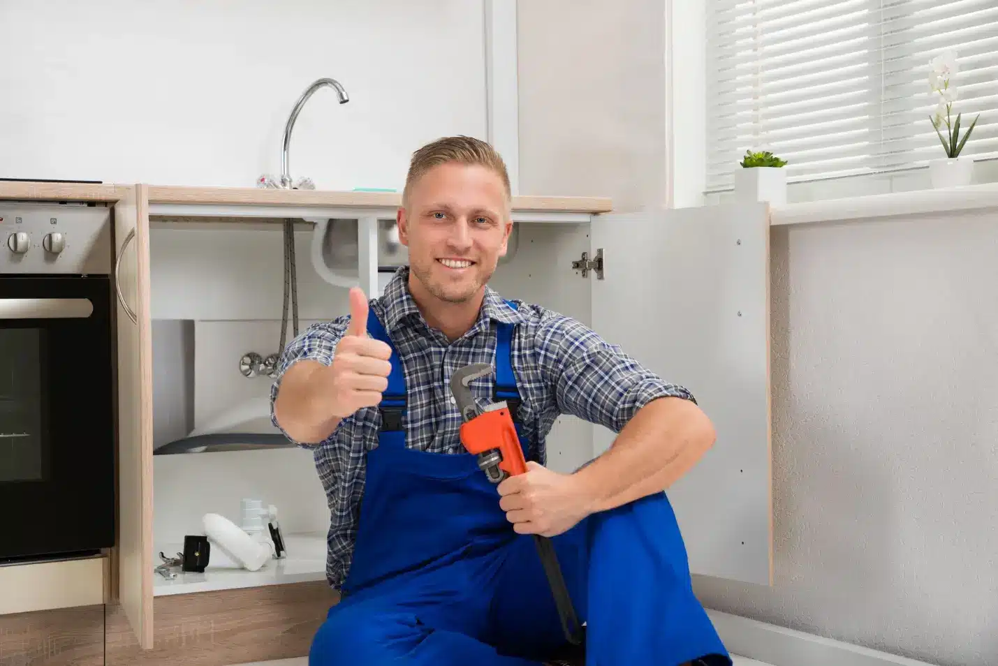 Plumber in blue overalls holding wrench giving thumbs up under kitchen sink