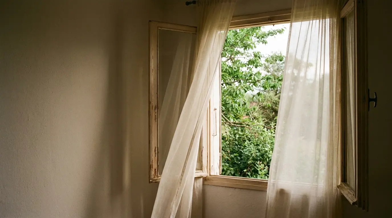 Open window with sheer curtains blowing in breeze revealing lush green trees outside