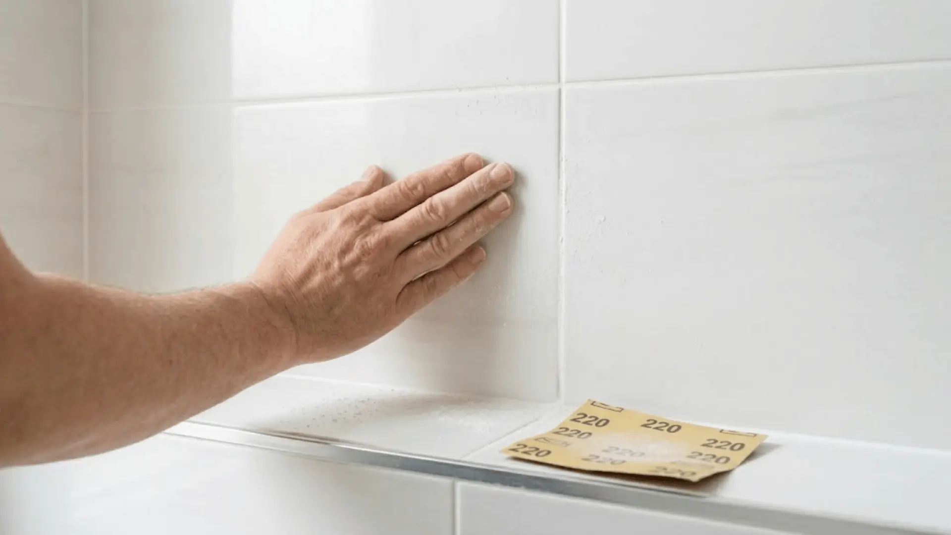 hand sanding a white ceramic wall tile with fine sandpaper to prepare surface for painting