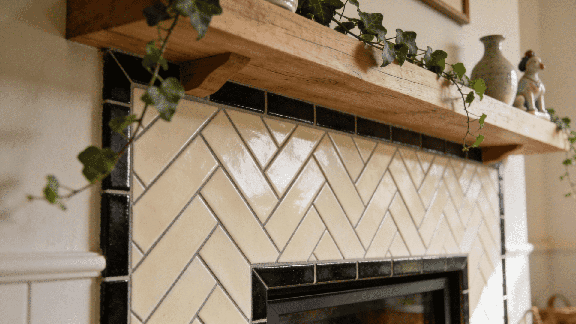 _a close up of a fireplace surround tiled in cream glazed herringbone tiles with a black gloss border trim beneath a chunky natural oak mantel decorated with trailing ivy and ceramic pieces