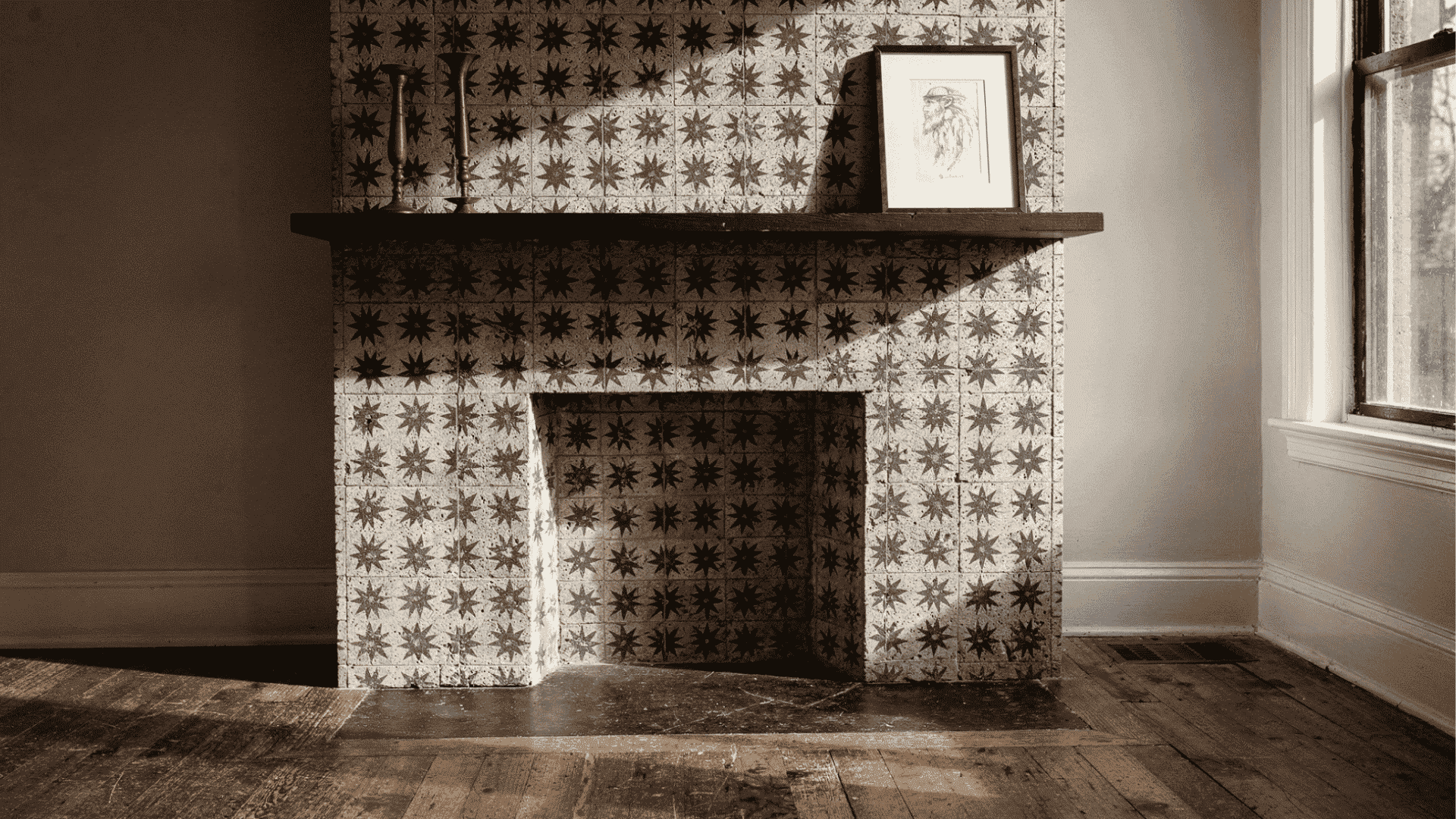 _a full fireplace surround covered entirely in repeating encaustic cement star pattern tiles in aged cream and charcoal with a flat dark wood mantel shelf and a single framed print leaned against