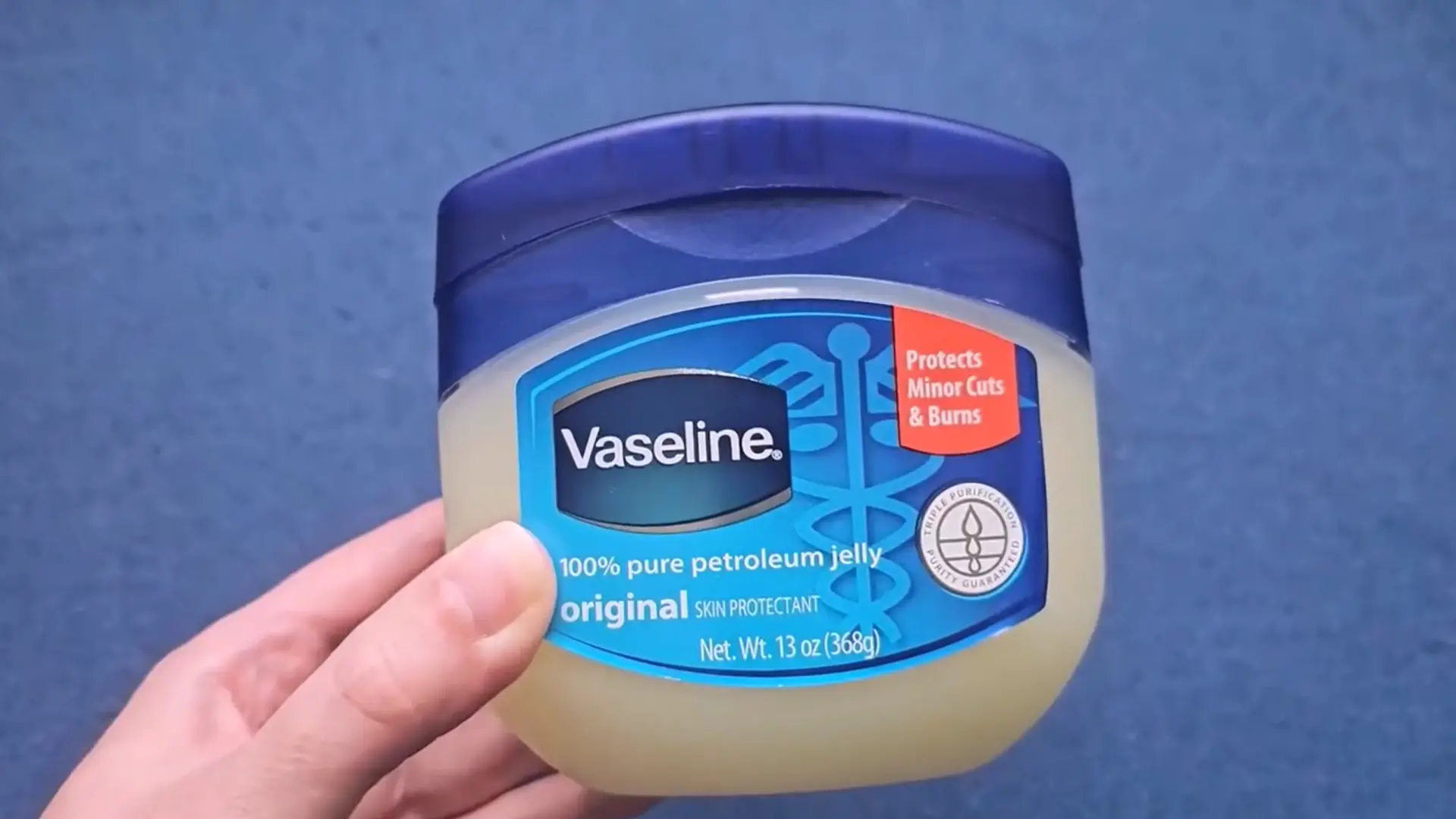 a hand holding a jar of vaseline petroleum jelly against a plain dark blue background wall