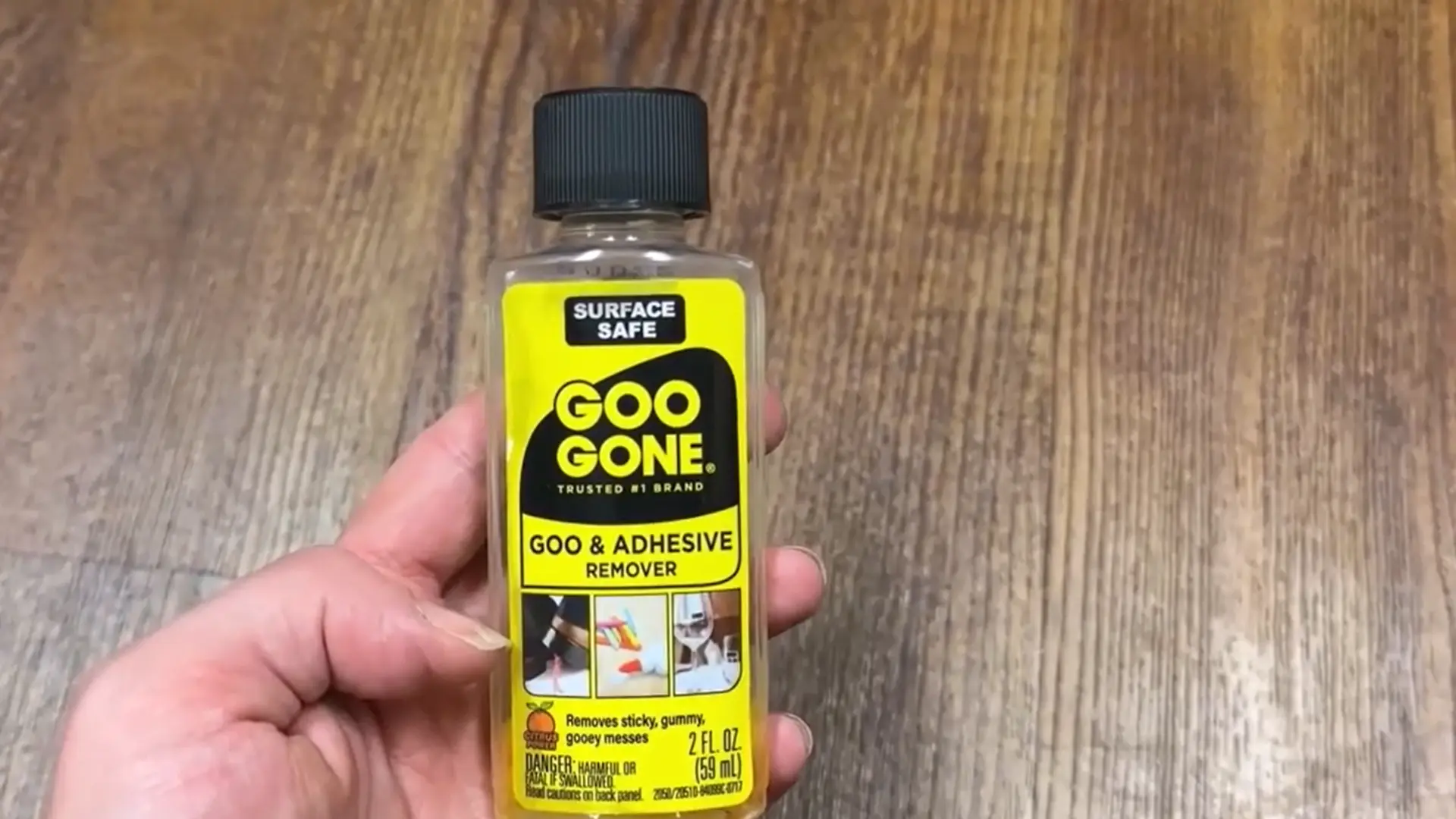 a hand holding a small bottle of goo gone adhesive remover over a brown wood floor surface