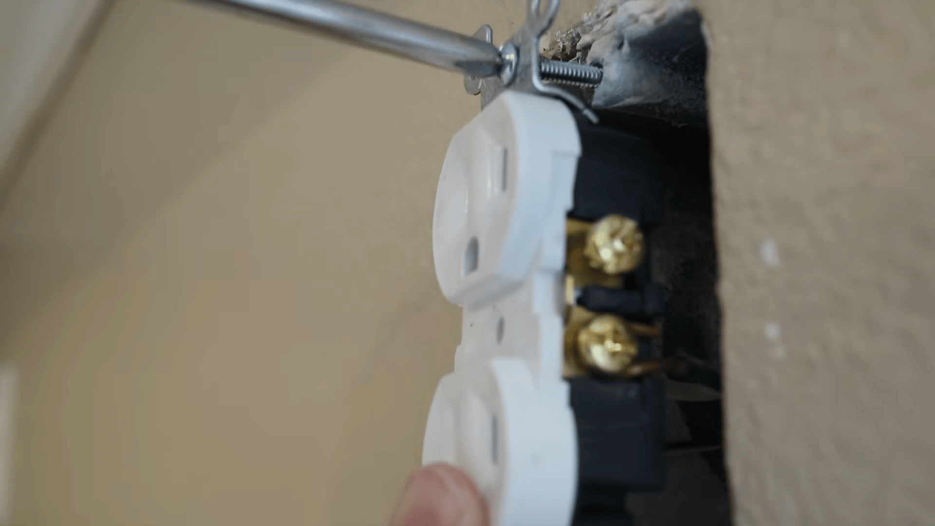 a hand using a screwdriver to screw on a new power outlet hanging onto a beige wall