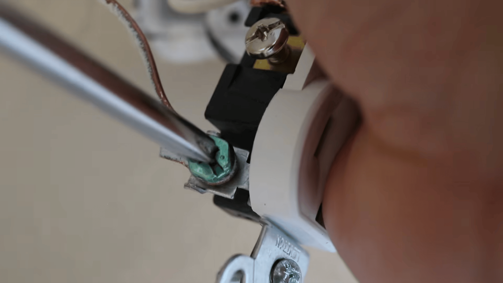 a hand using a screwdriver to screw the green bolt connected to a bare wire of a power outlet hanging from a beige wall