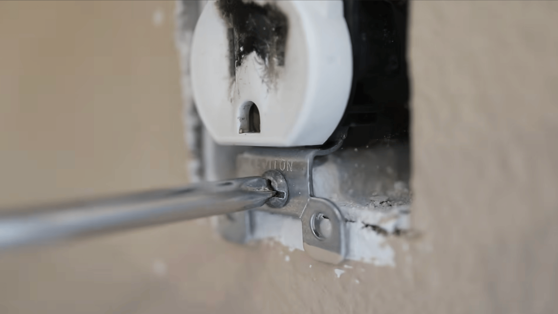 a hand using a screwdriver to unscrew the bottom of a white power outlet on a beige wall