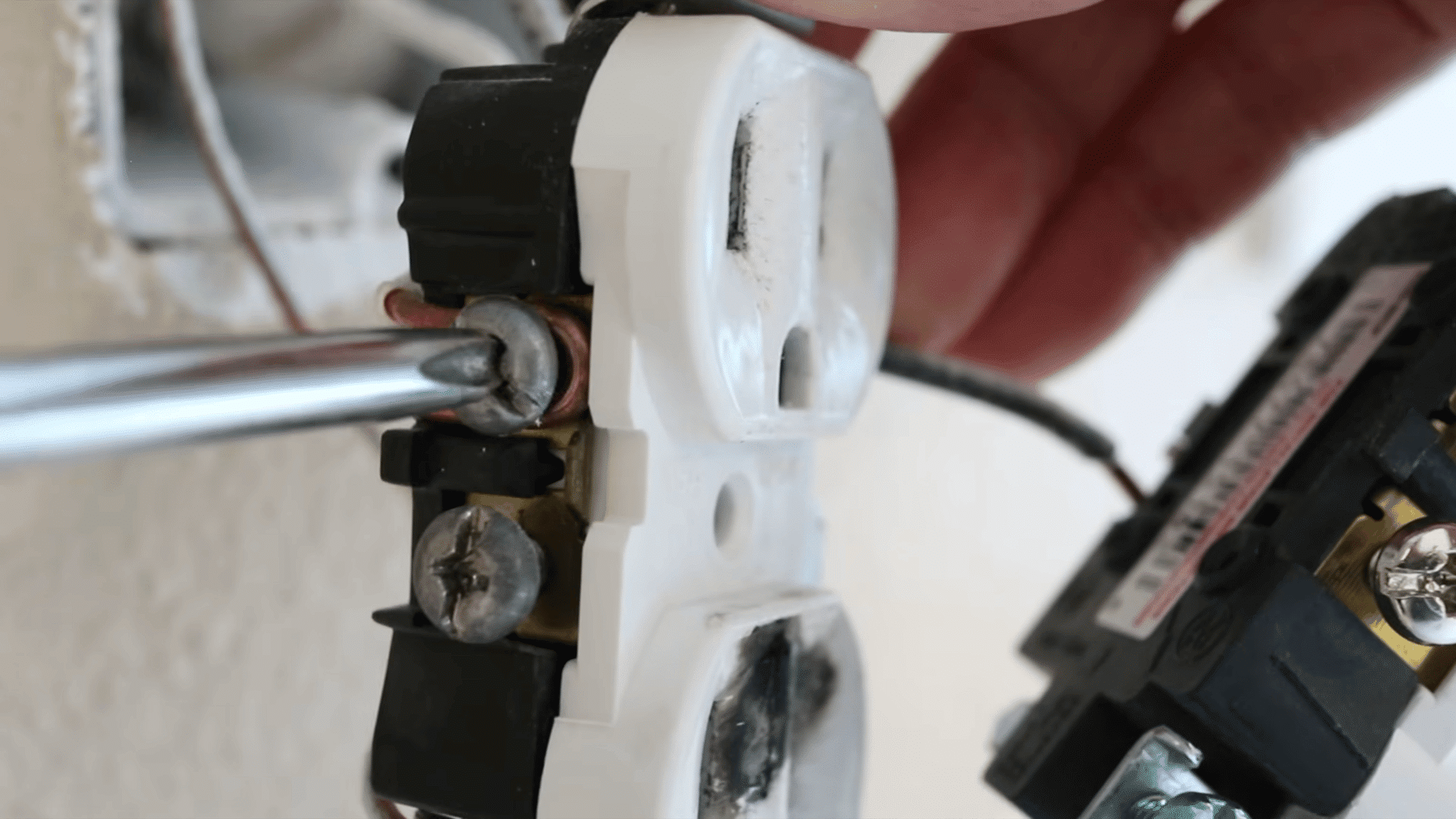 a hand using a screwdriver to unscrew the golden screw connected to the a white wire of a power outlet hanging from a beige wall