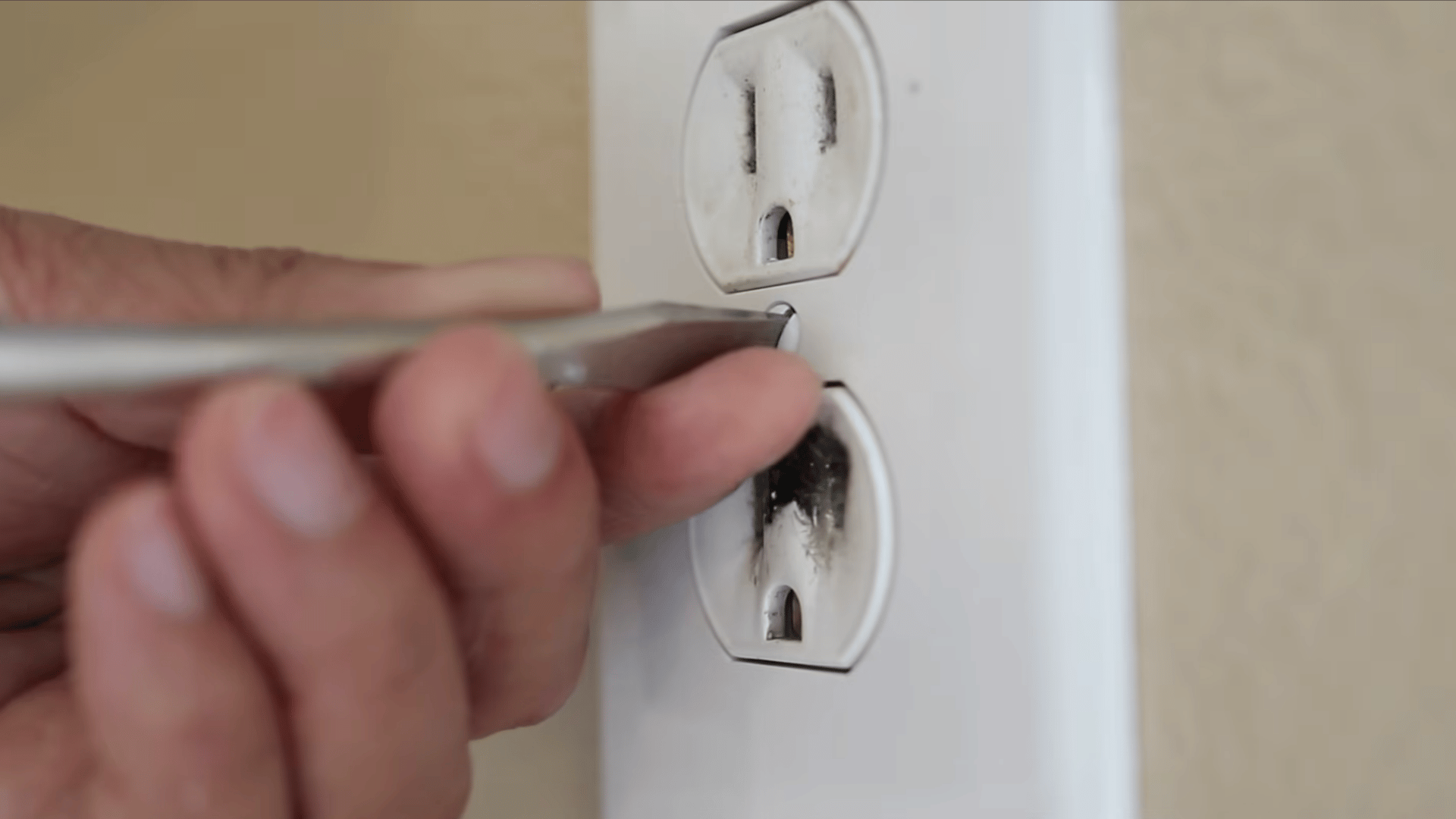 a hand using a screwdriver unscrewing thebolt of a burnt white power outlet on a beige wall