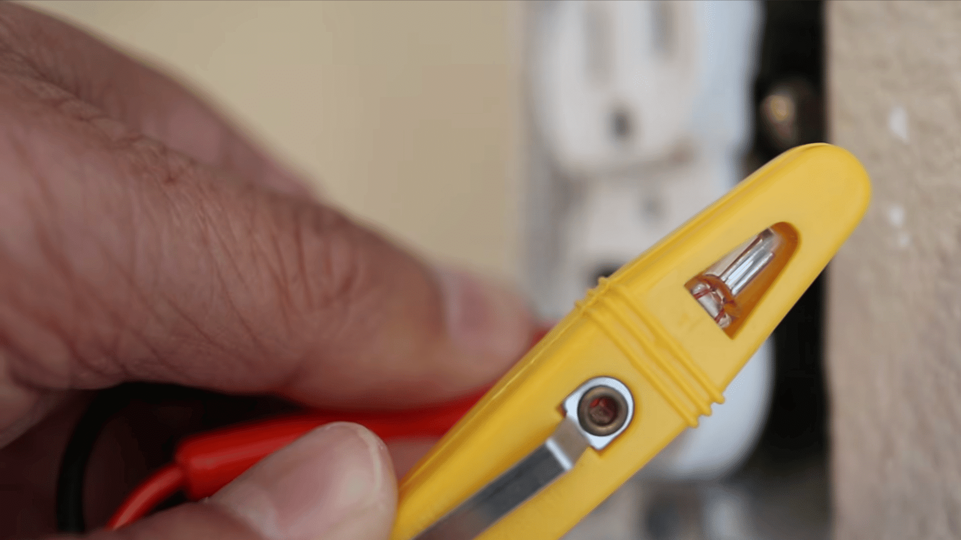 a hand using a small yellow voltage tester on a white power outlet on a beige wall