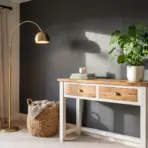 A living room corner featuring a white and wood console table with a Monstera plant, a brass arc floor lamp, and a woven storage basket against a dark gray accent wall