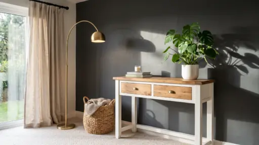 A living room corner featuring a white and wood console table with a Monstera plant, a brass arc floor lamp, and a woven storage basket against a dark gray accent wall