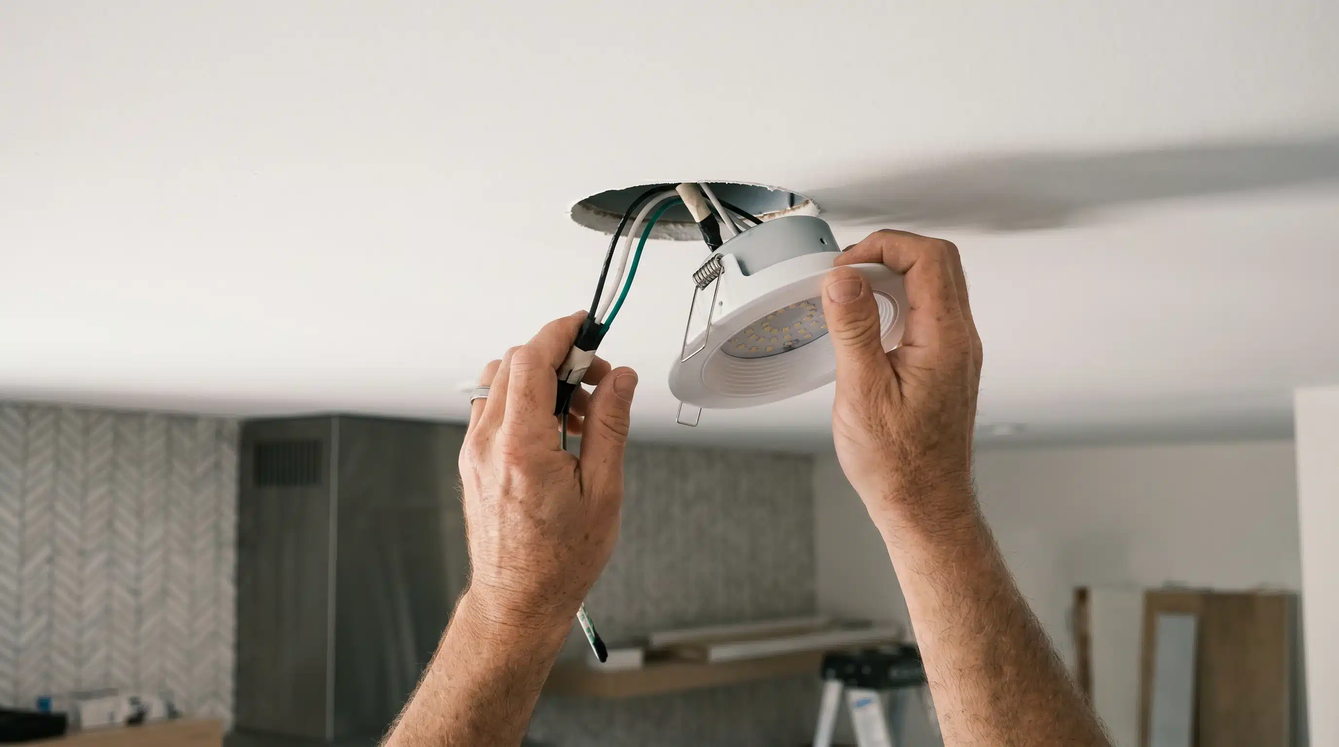 A man is installing a ceiling light fixture in a bright room, focused on his task while on a ladder