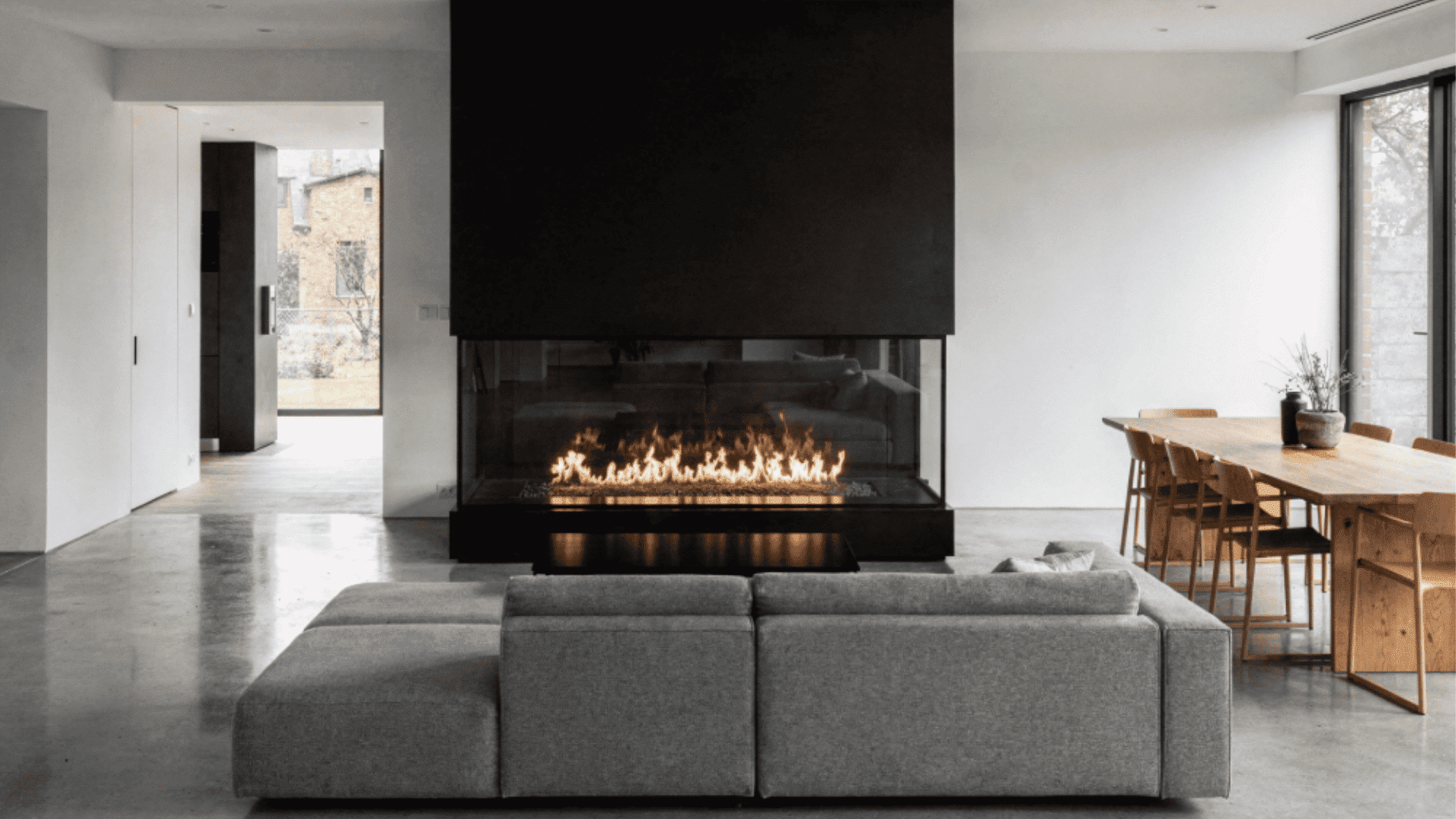 a modern living room with a large black three sided fireplace and a gray sectional sofa in the foreground and a wooden dining table with chairs in the background